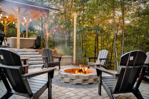 An outdoor patio with Adirondack chairs, a stone fire pit, and a covered seating area with string lights.