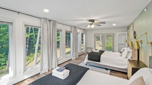 A modern bedroom with two beds, large windows, a ceiling fan, and a view of the forest.