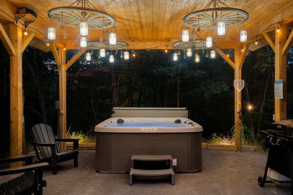 A hot tub under a wooden pergola with string lights, surrounded by Adirondack chairs and a grill.