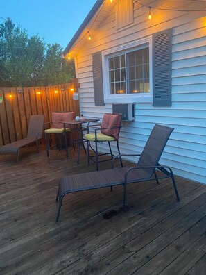 Una terraza de madera con muebles de exterior, incluyendo sillas y una mesa, y luces colgantes.