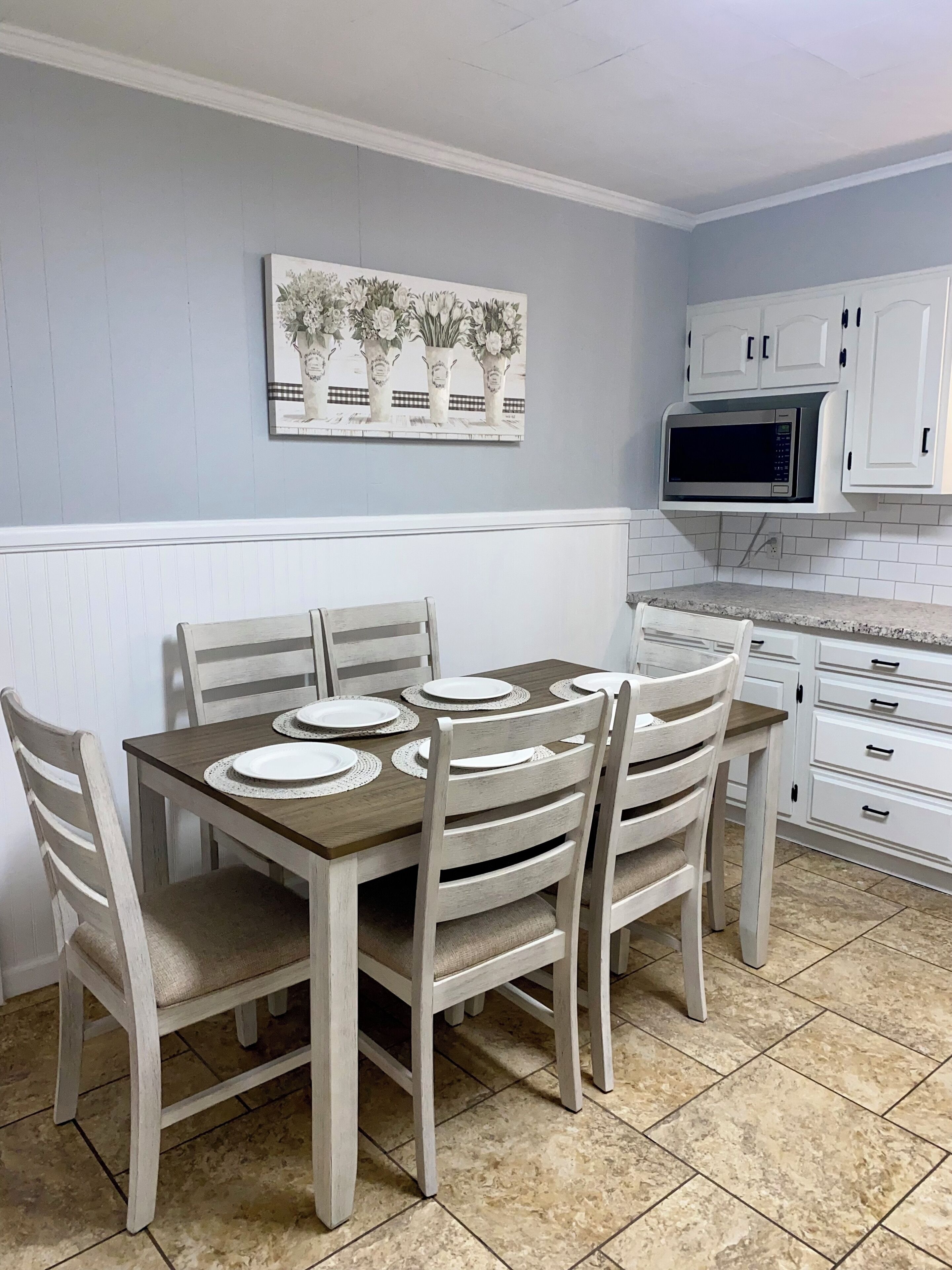 Comedor con mesa de madera dispuesta para cuatro, sillas blancas y un mueble blanco con un microondas encima.