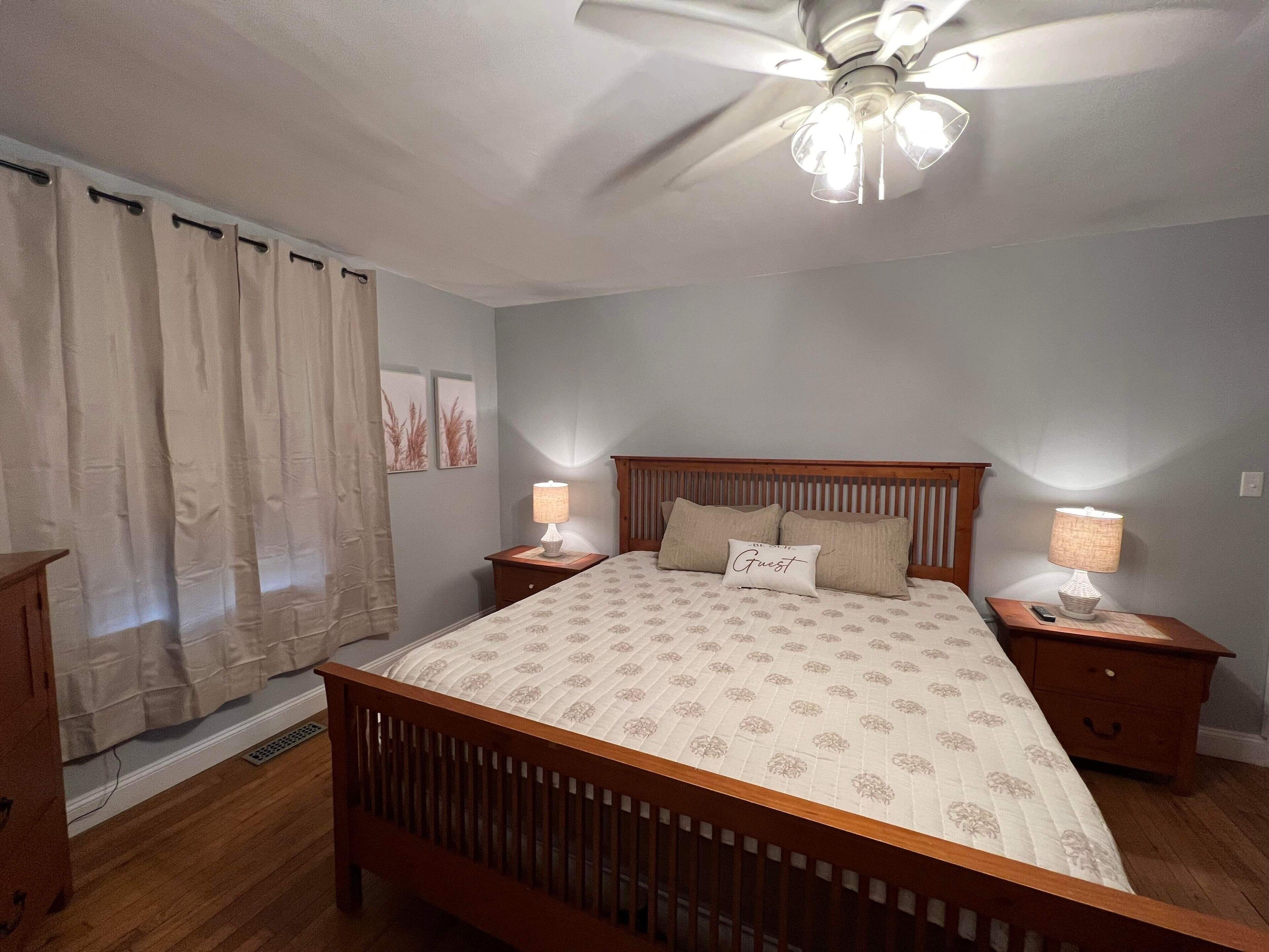 A bedroom with a wooden bed, two bedside tables with lamps, a ceiling fan, and a window with curtains.