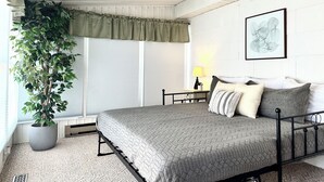 Ein Schlafzimmer mit einem Bett, einer Topfpflanze, einem Fenster mit Jalousien, einem Nachttisch mit Lampe und einem gerahmten Bild an der Wand.