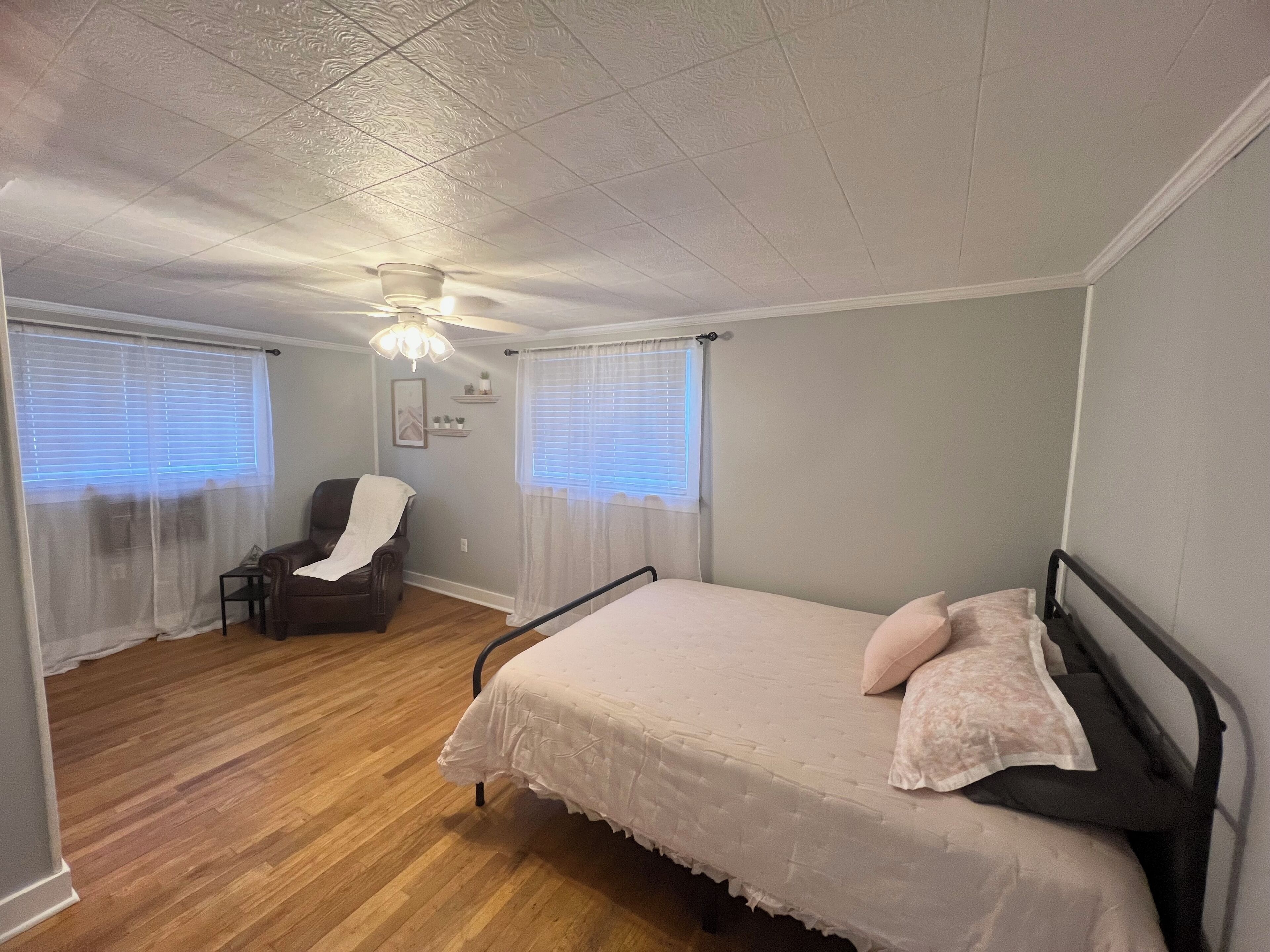 Ein Schlafzimmer mit einem Bett, einem Sessel, einem Deckenventilator und zwei Fenstern mit Jalousien.