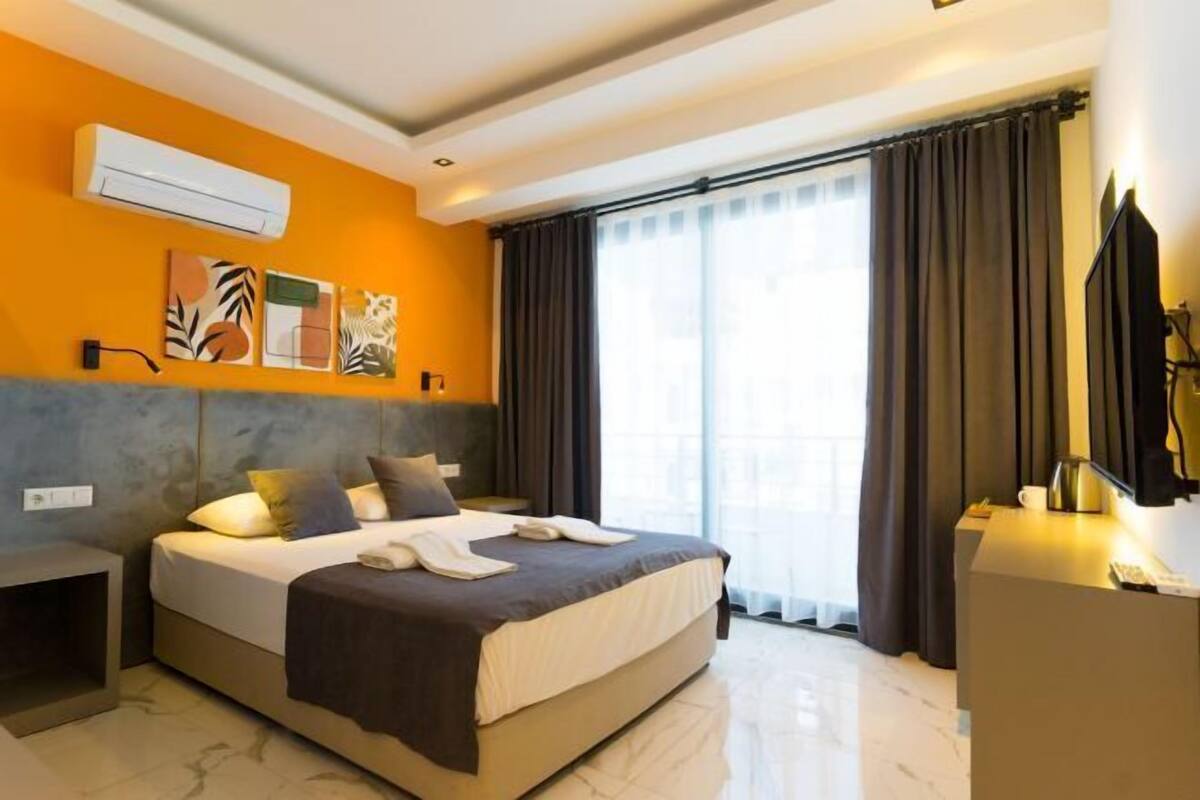 A modern hotel room with a bed, a desk, an air conditioning unit, artwork on the wall, and a large window with curtains.