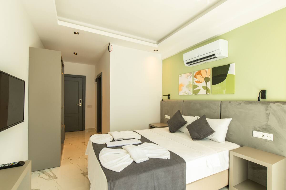 A modern hotel room with a large bed, a flat-screen TV, an air conditioning unit, and a small bedside table.