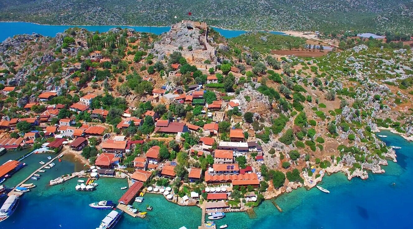 Vista aérea de un pueblo costero con casas, una estructura fortificada y un puerto deportivo con barcos.
