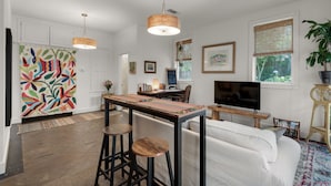 Uma sala de estar moderna com um sofá branco, uma mesa de centro de madeira e uma TV. Há duas luminárias pendentes, um quadro abstrato colorido e uma janela com persianas.