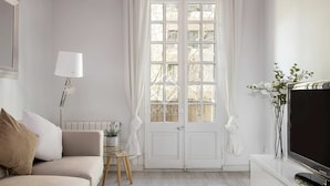 Un salon minimaliste comprenant un canapé, une petite table d’appoint, une lampe, un téléviseur et une grande fenêtre avec des rideaux.