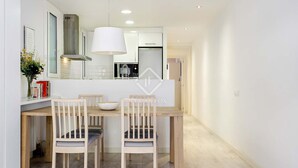Une cuisine moderne avec des meubles blancs, un grand luminaire suspendu blanc et un coin repas composé de deux chaises en bois et d’une table blanche.