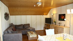Una sala de estar con techo de madera, un sofá gris, una mesa de centro, un televisor y una zona de comedor con mesa y sillas.