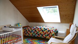 Una habitación acogedora con techo inclinado, una cama con colcha colorida, una cuna blanca, una silla de madera y un ventanal pequeño.