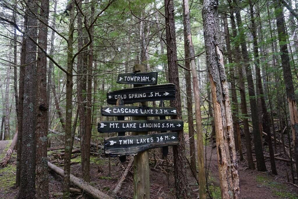 Ein Wegweiser in einem bewaldeten Gebiet mit mehreren Pfeilen, die zu verschiedenen Zielen zeigen, darunter 'TOWER 1M', 'COLD SPRING 5M', 'CASCADE LAKE 2.3M', 'MT. LAKE LANDING 5.5M' und 'TWIN LAKES 34M'.