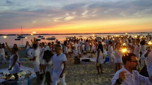 A beach with people holding sparklers, boats in the water, and a sunset in the background.