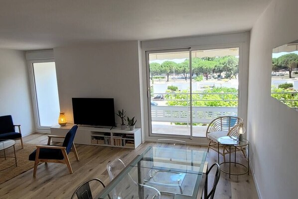A modern living room with a flat-screen TV, a glass dining table, and a large sliding glass door leading to a balcony with a view of greenery and a parking area.