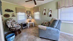 A living room with a ceiling fan, a blue sofa, a patterned armchair, a small table with a lamp, and decorative wall art. - Cozy Cottage in quiet neighborhood not far from beaches (Marshallberg)