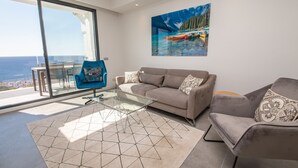 Ein modernes Wohnzimmer mit einer Couch, Sesseln und einem Glastisch. Ein großes Fenster bietet Blick auf das Meer und die Berge.