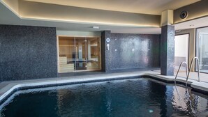 Ein modernes Hallenbad mit Glaswand, einem Holz-Saunabereich und einer Metallleiter, die in das Becken führt.