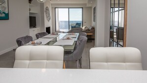 Ein zeitgemäßer Essbereich mit einem langen Tisch, weißen gepolsterten Stühlen und Blick auf das Meer durch große Fenster.