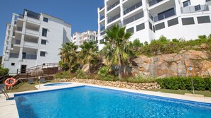 A swimming pool with a stone border and a lifebuoy, surrounded by a white building with balconies and palm trees.