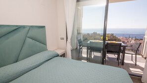A balcony with a table and chairs, a bed with a green headboard, and a view of the sea and buildings.
