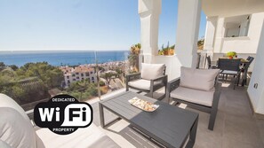 A balcony with a view of the sea and a cityscape, furnished with a table and chairs, and a WiFi symbol indicating connectivity.