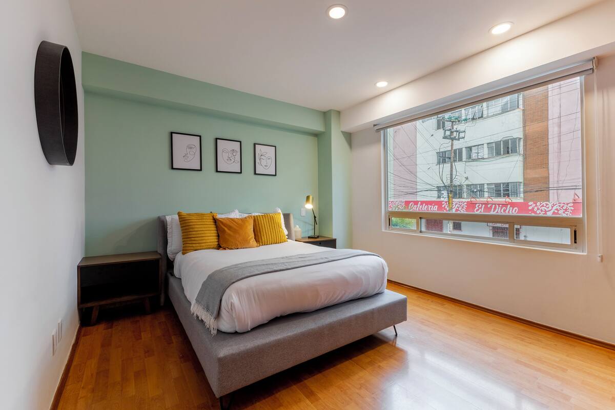 A bedroom with a bed, bedside tables, a window with a view of buildings, and framed artwork on the wall.