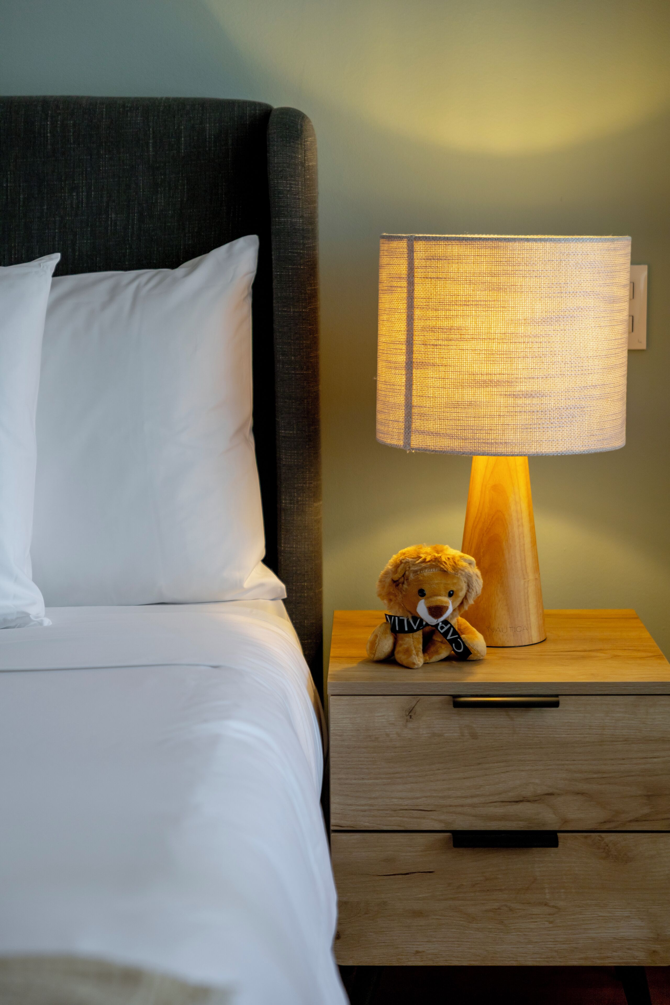 Una cama con ropa de cama blanca, un cabecero oscuro, una mesita de noche de madera con una lámpara y un pequeño peluche.