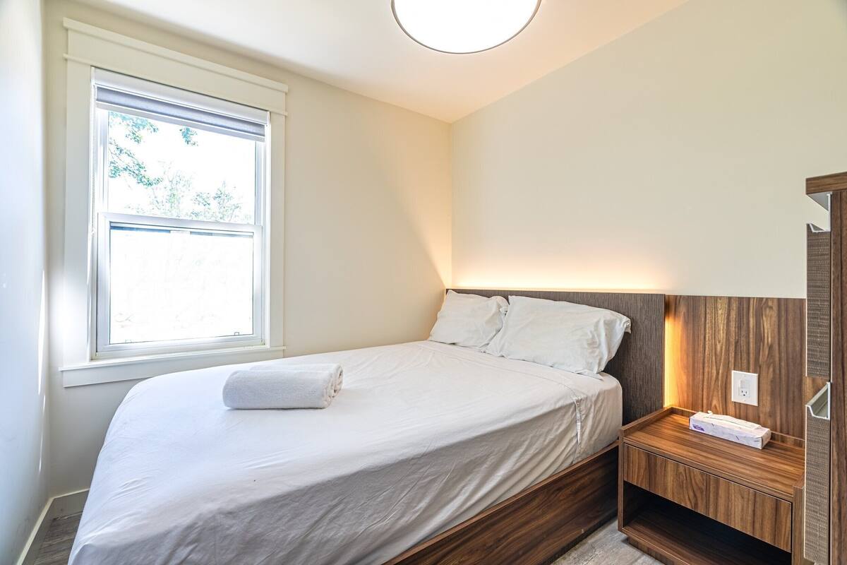 A bedroom with a bed, a nightstand, a window, and a wooden headboard.