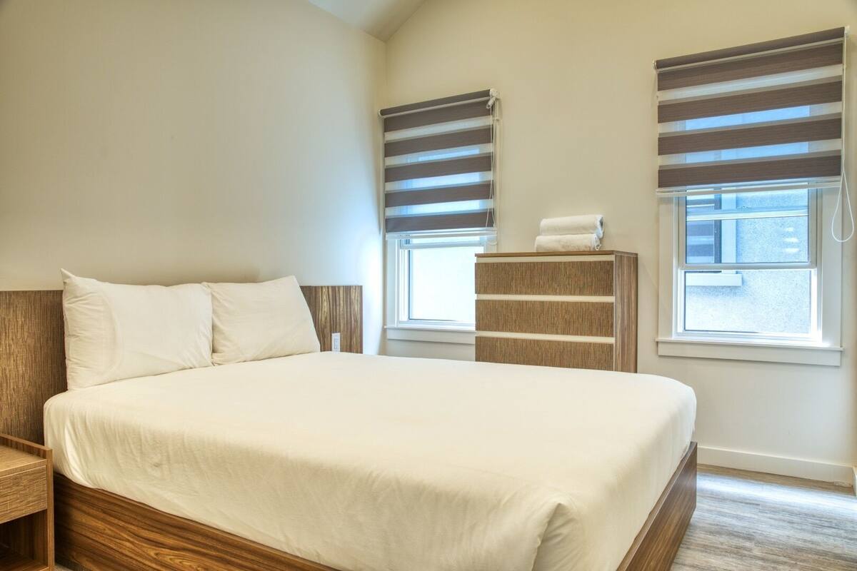 A bedroom with a large bed, two bedside tables, a wooden dresser, and two windows with blinds.