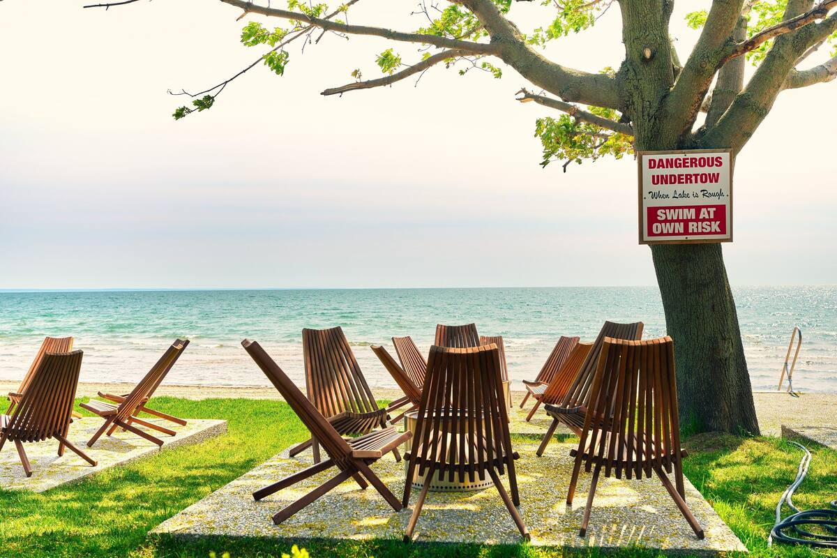 A beachside setting with wooden lounge chairs arranged under a tree, a sign warning of dangerous undertow, and a view of the ocean.