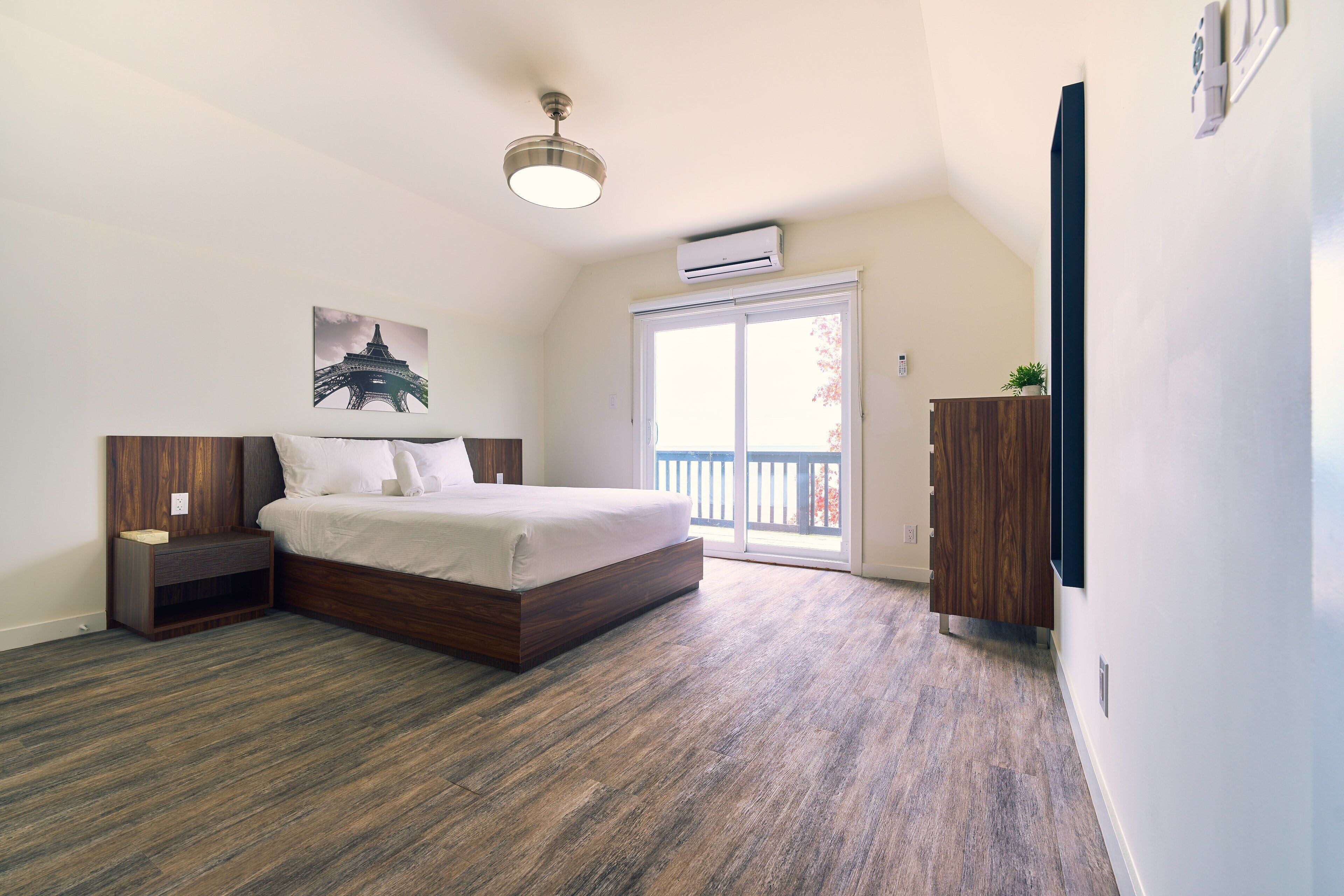A bedroom with a wooden bed, a nightstand, a wall-mounted air conditioner, and a large window with a view of a balcony.