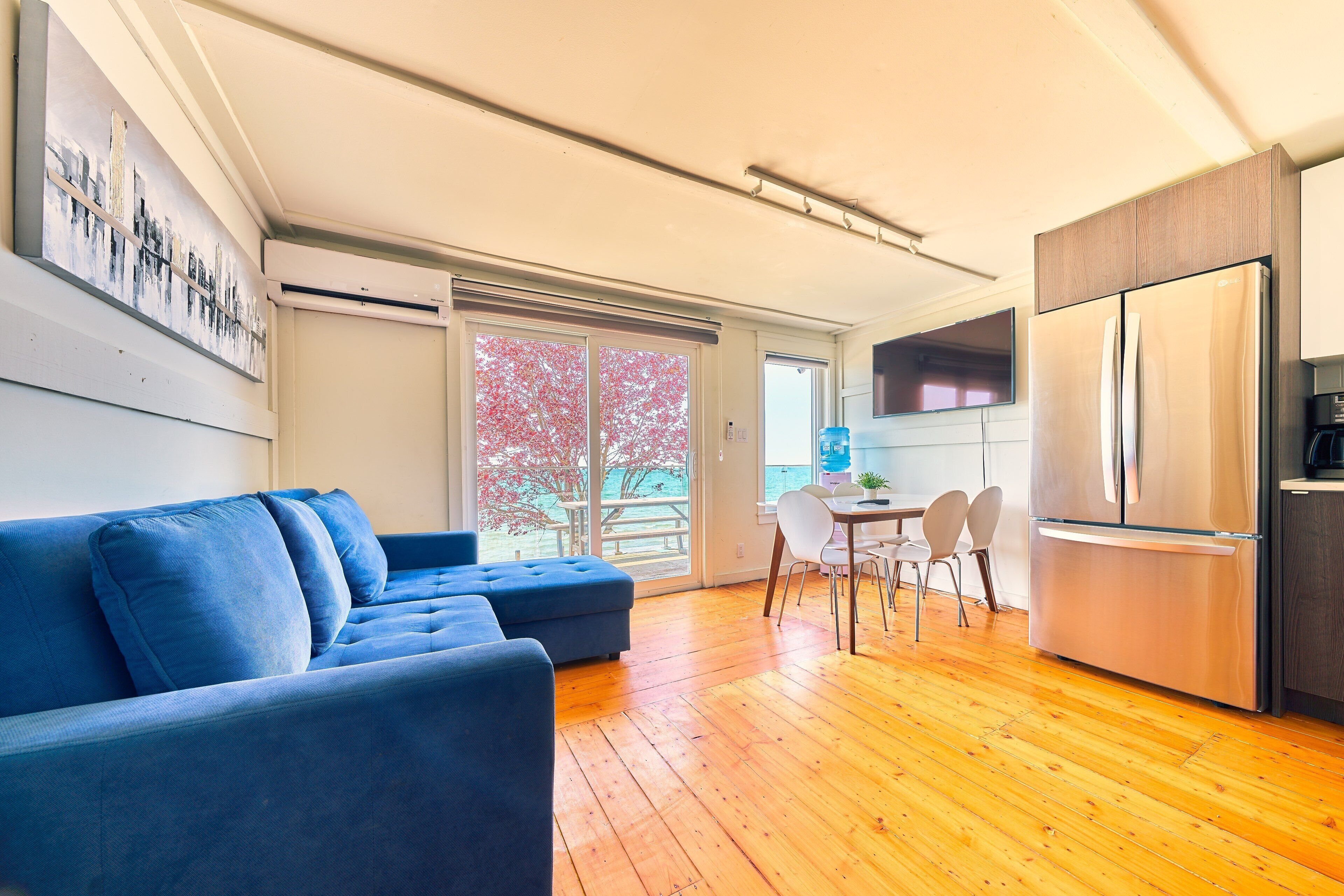 Una sala de estar moderna con un sofá azul, piso de madera, una cocina con refrigerador de acero inoxidable y un comedor con mesa y sillas.