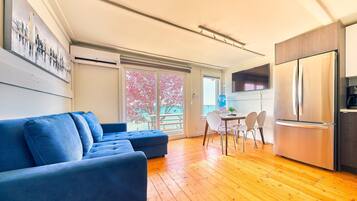 Una sala de estar moderna con un sofá azul, piso de madera, una cocina con refrigerador de acero inoxidable y un comedor con mesa y sillas.