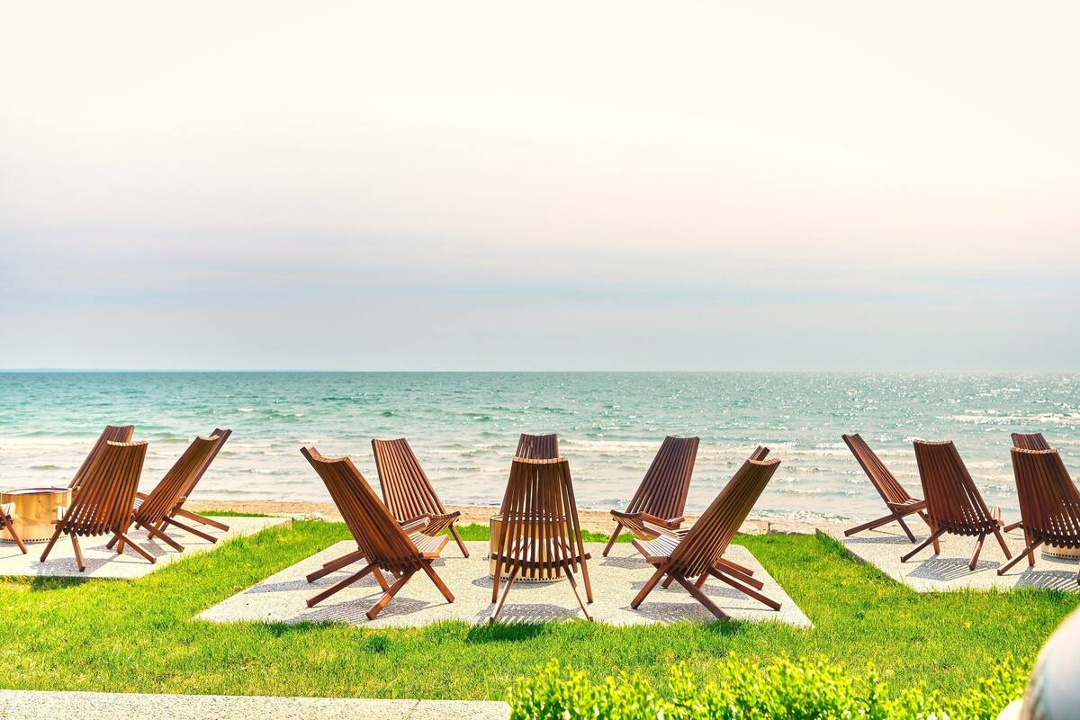 A row of wooden lounge chairs facing the ocean, arranged on a concrete platform with a grassy area in the foreground.