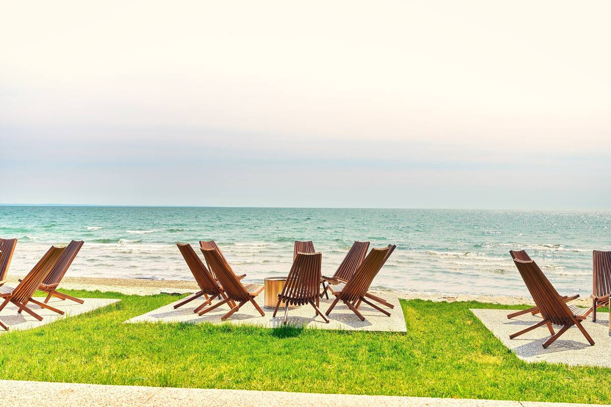 A row of wooden lounge chairs and a fire pit on a grassy area by the ocean.