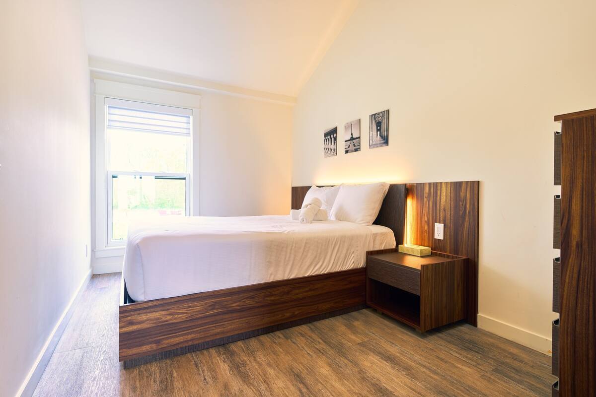 A modern bedroom with a large bed, a wooden nightstand, and a window with blinds.