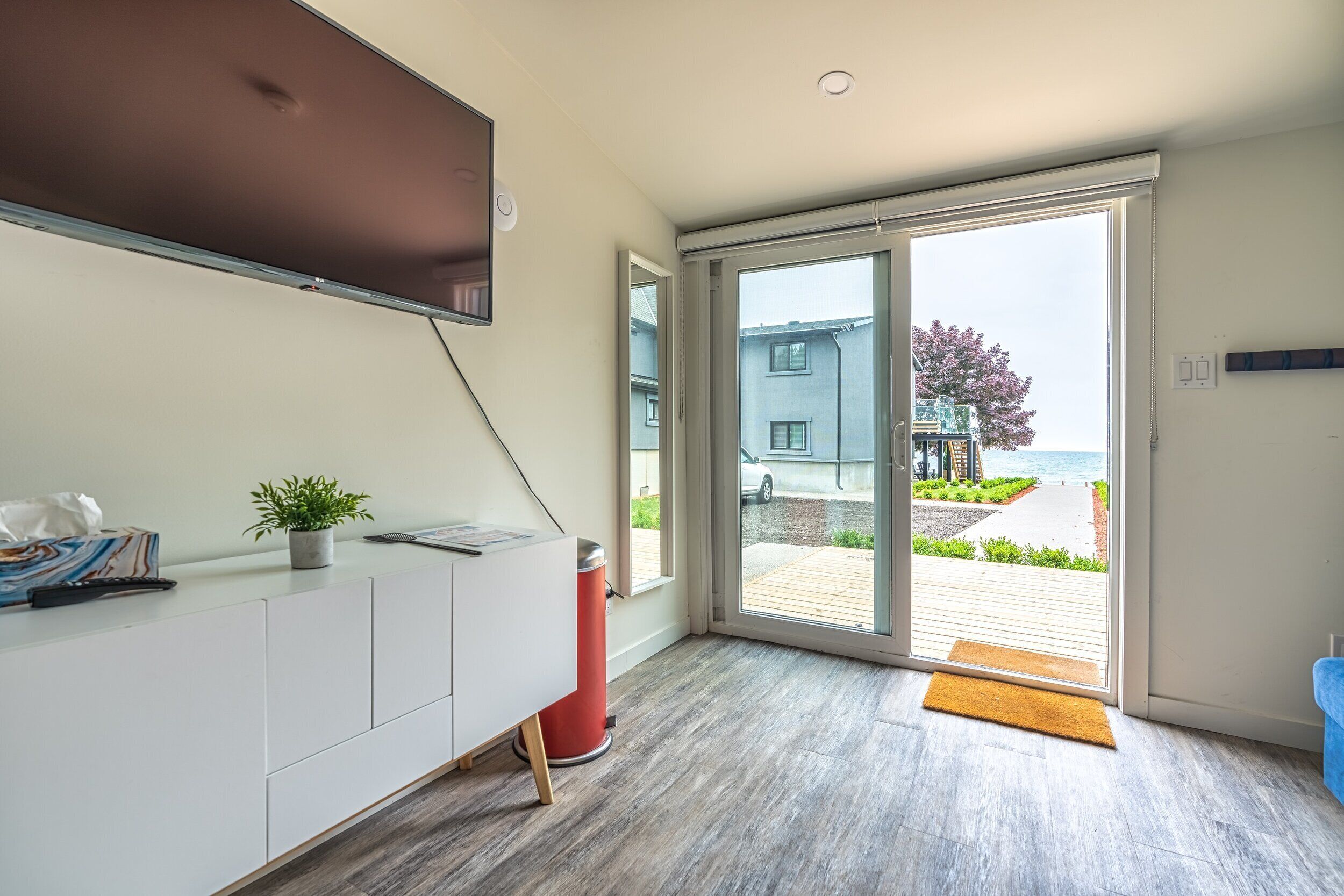Una habitación moderna con un amplio ventanal corredera que da a un espacio exterior, un armario blanco con una planta y un televisor de pantalla plana montado en la pared.