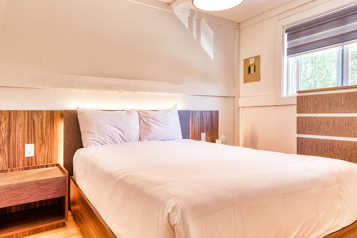 A neatly made bed with a wooden headboard, a bedside table, and a window with blinds.