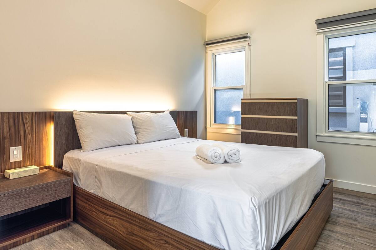 A neatly made bed with a wooden headboard, a bedside table with a lamp, and a window with blinds.