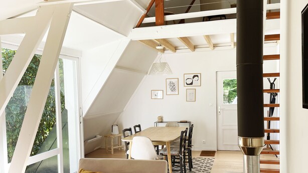 A modern loft with a high ceiling, exposed wooden beams, a central fireplace, and a dining area with a table and chairs.