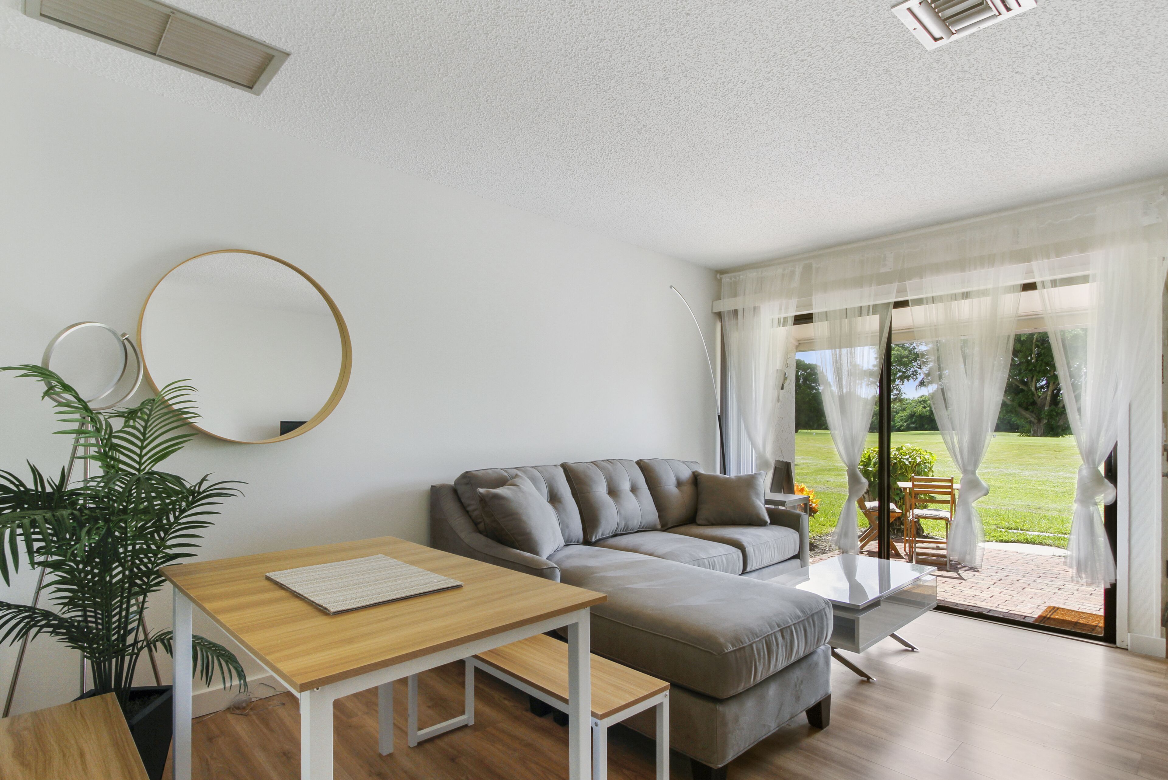 Ein modernes Wohnzimmer mit einem grauen Sofa, einem Holztisch, einer Topfpflanze und einem großen Fenster mit transparenten Vorhängen.