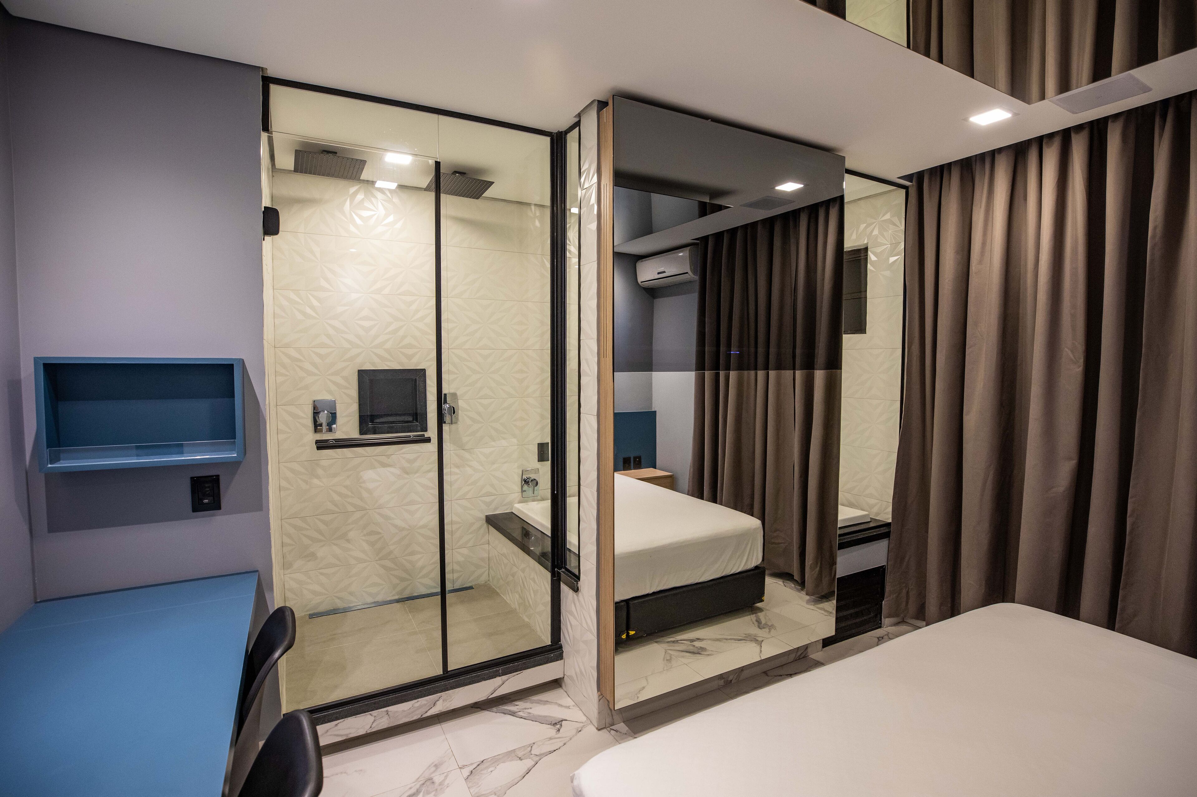 A modern hotel room with a glass shower enclosure, a bed, a bedside table, and a built-in TV shelf.