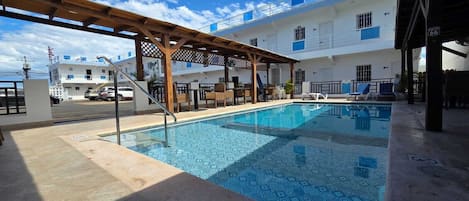A swimming pool with a wooden pergola, lounge chairs, and a building with balconies in the background.