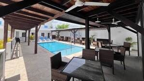 Un espace extérieur avec une piscine, des chaises longues et des tables, un ventilateur de plafond, et un bâtiment en arrière-plan.