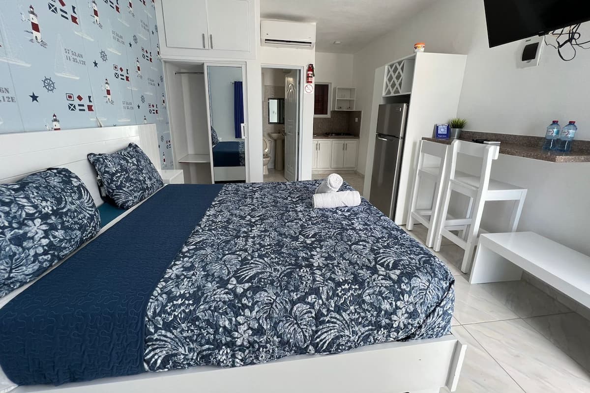 A bedroom with a bed, a desk, a chair, a TV, and a kitchenette.