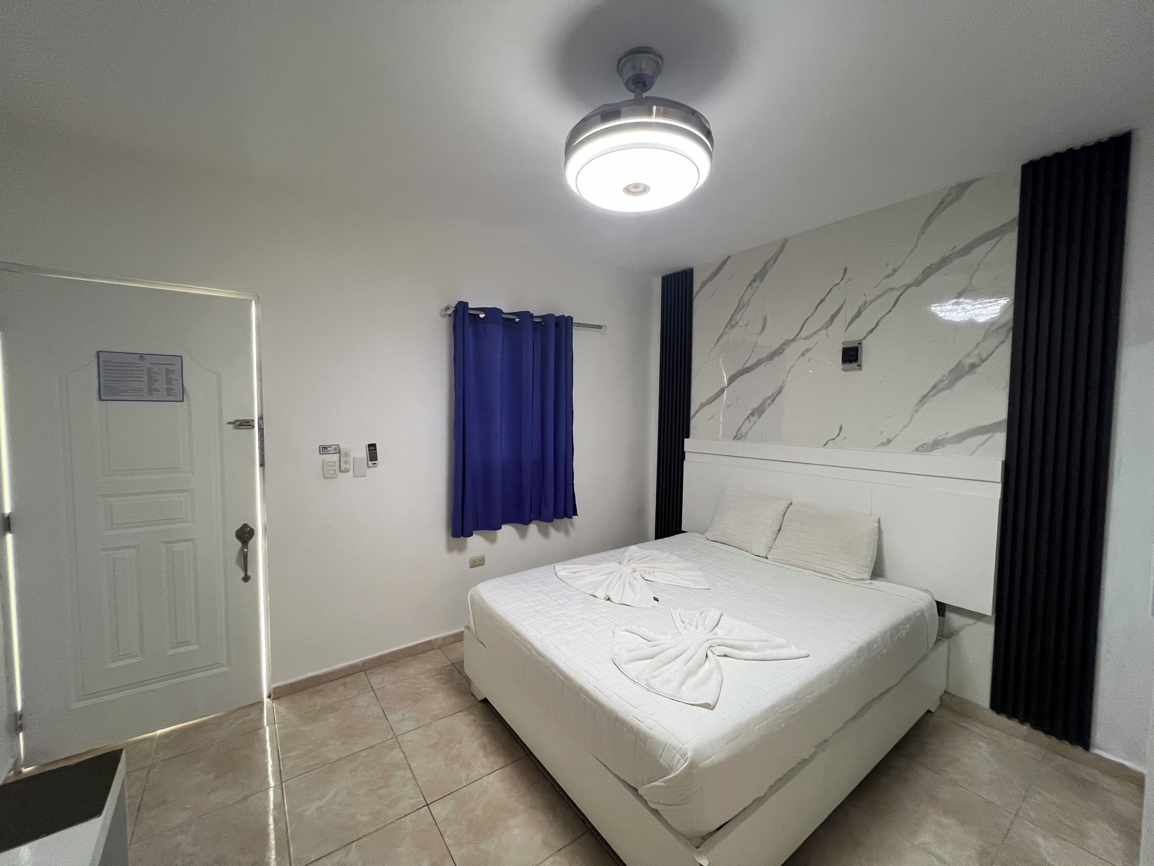 A hotel room with a bed, a ceiling light, a door, a wall with a marble-like pattern, and a blue curtain.
