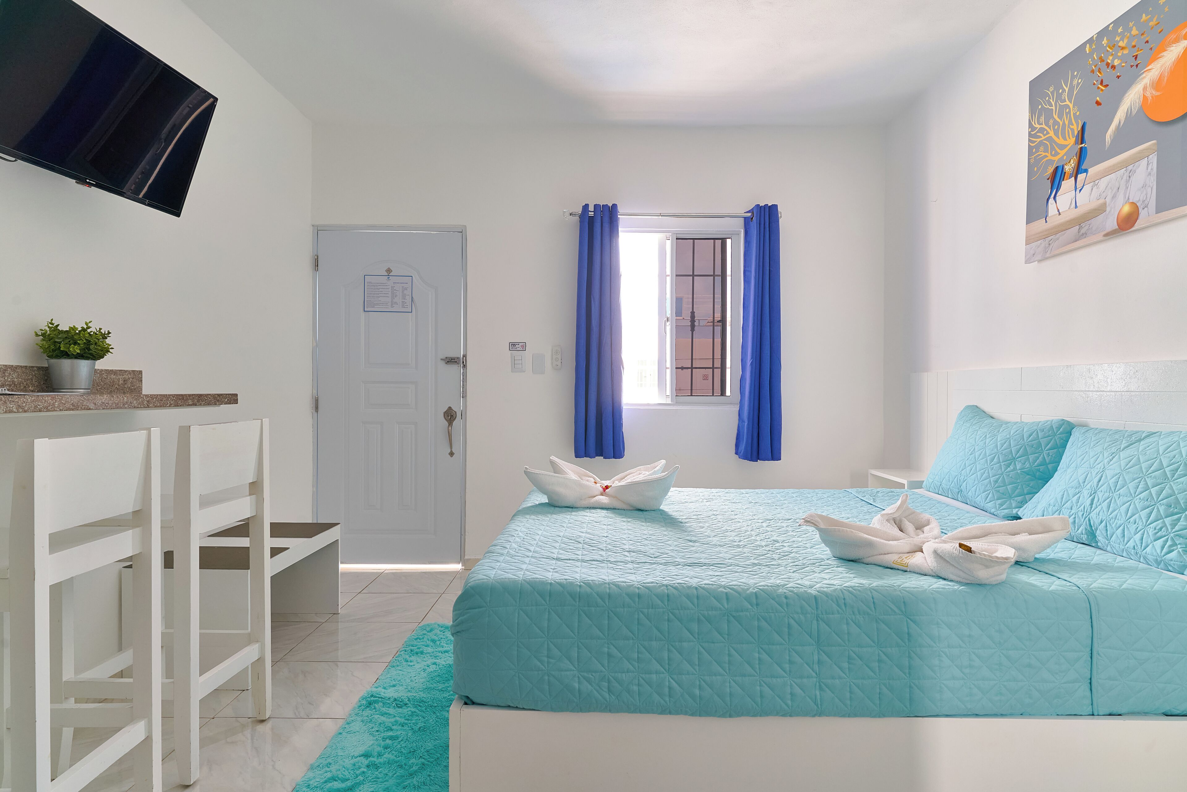 Un dormitorio ordenado con cama, escritorio, silla, televisor y ventana con cortinas.