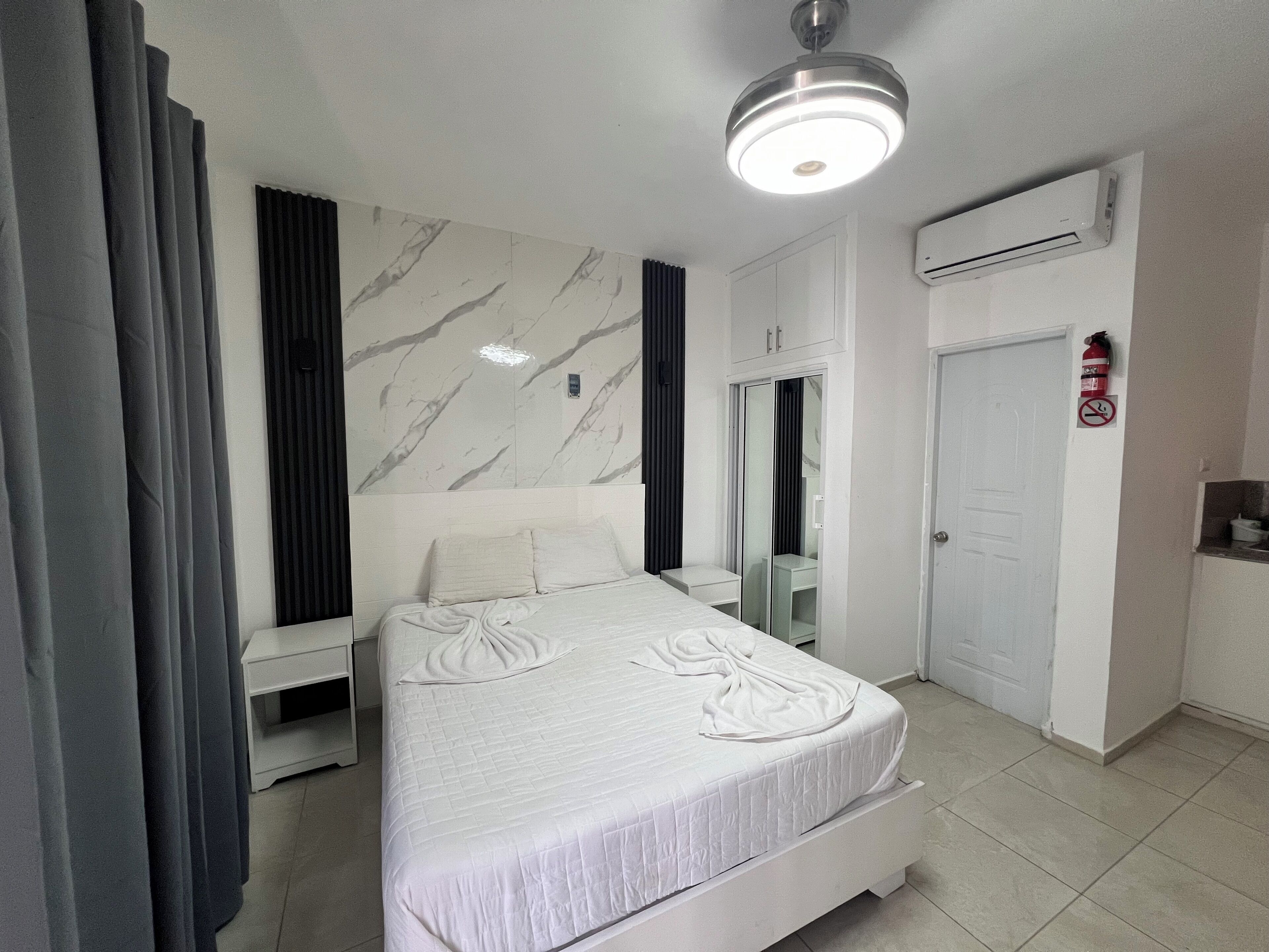 A modern hotel room with a marble-patterned wall, a bed with a neatly arranged white bedspread, a bedside table, a wardrobe, and an air conditioning unit.
