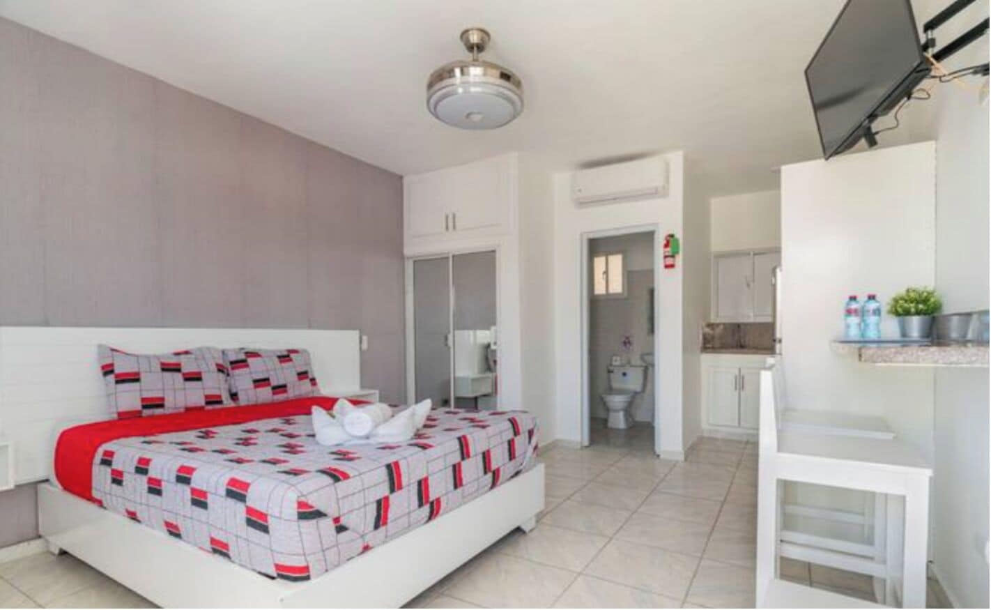 Un dormitorio moderno con una cama, ventilador de techo, televisor y un baño visible a través de una puerta abierta.